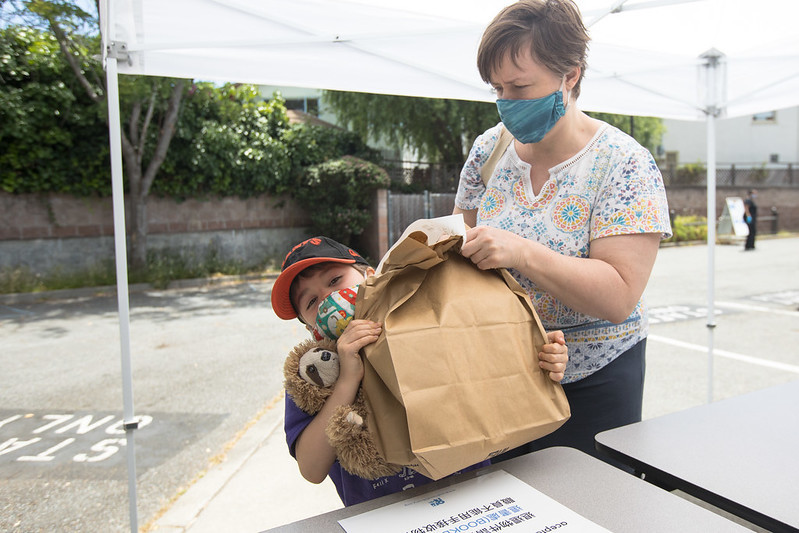 Woman with blue face masks helps young boy in orange hat take a big paper bag of books from the table outside of a library.