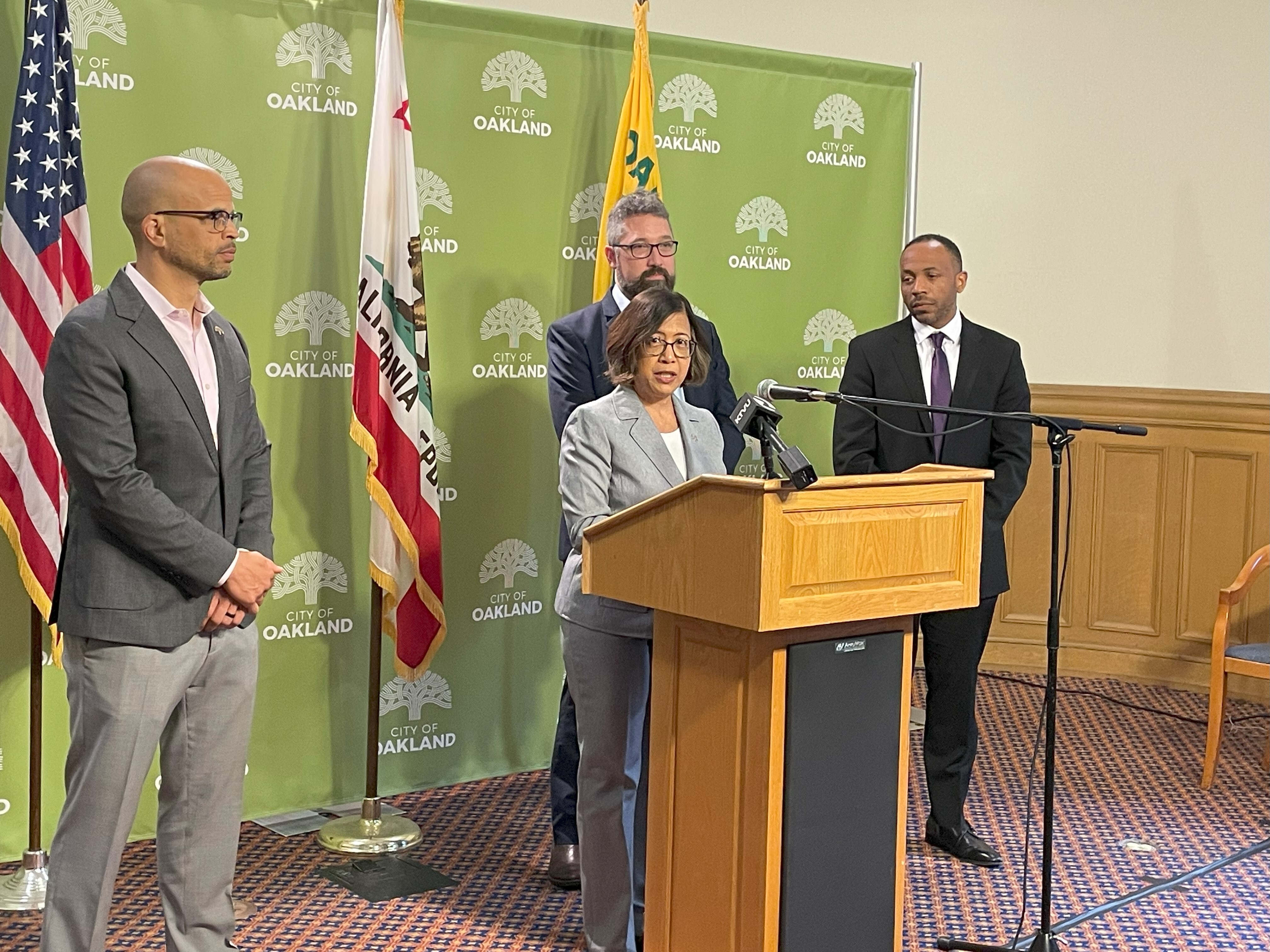 Four people standing, one behind a podium, green City of Oakland logo in the backgroud.