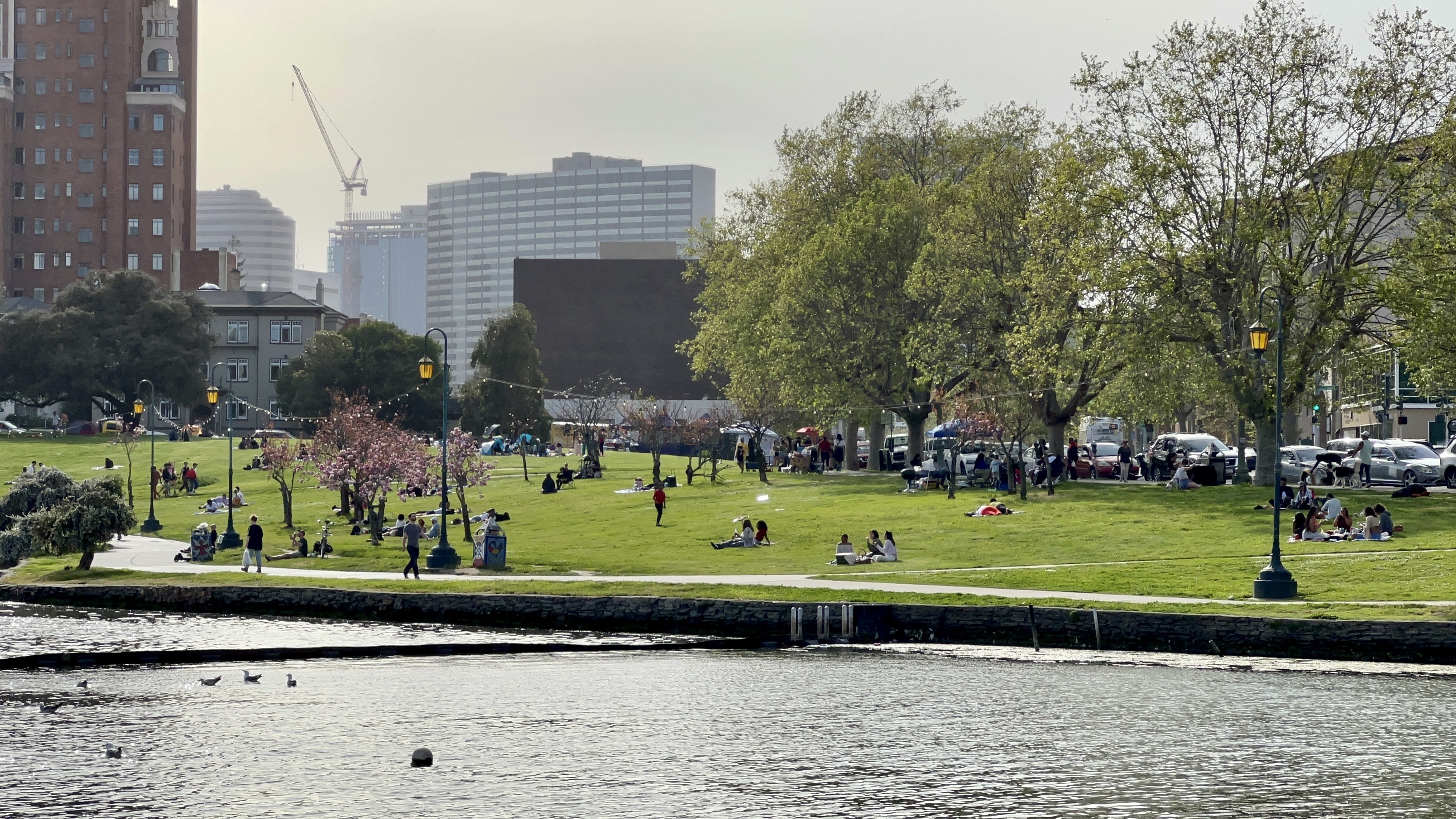 Distance shot of part of Lake Merritt, with buildings in the background, people sitting on law, walking on path on an overcast day.